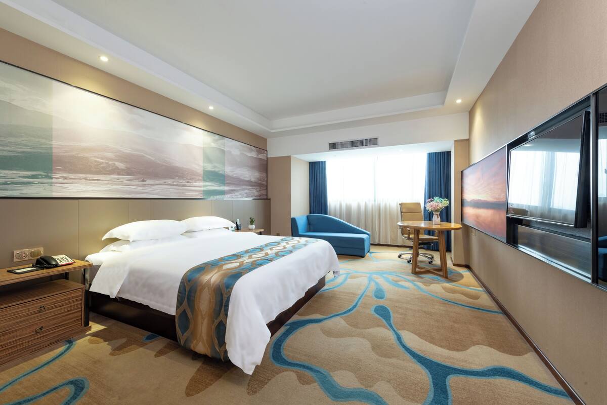 Habitación de hotel con una cama grande, un escritorio con teléfono, una silla, un televisor y un ventanal con cortinas.