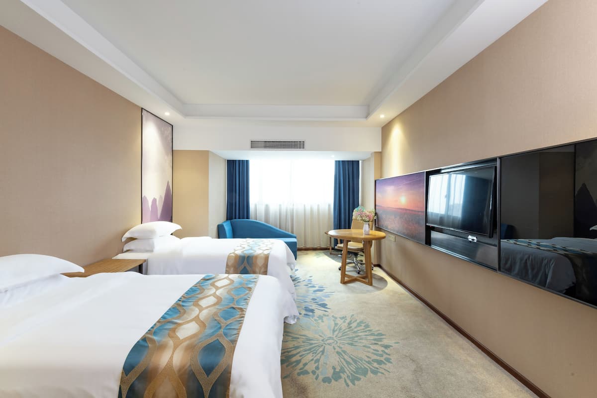 Habitación de hotel con dos camas, un escritorio, una silla, un televisor y un ventanal grande con cortinas.