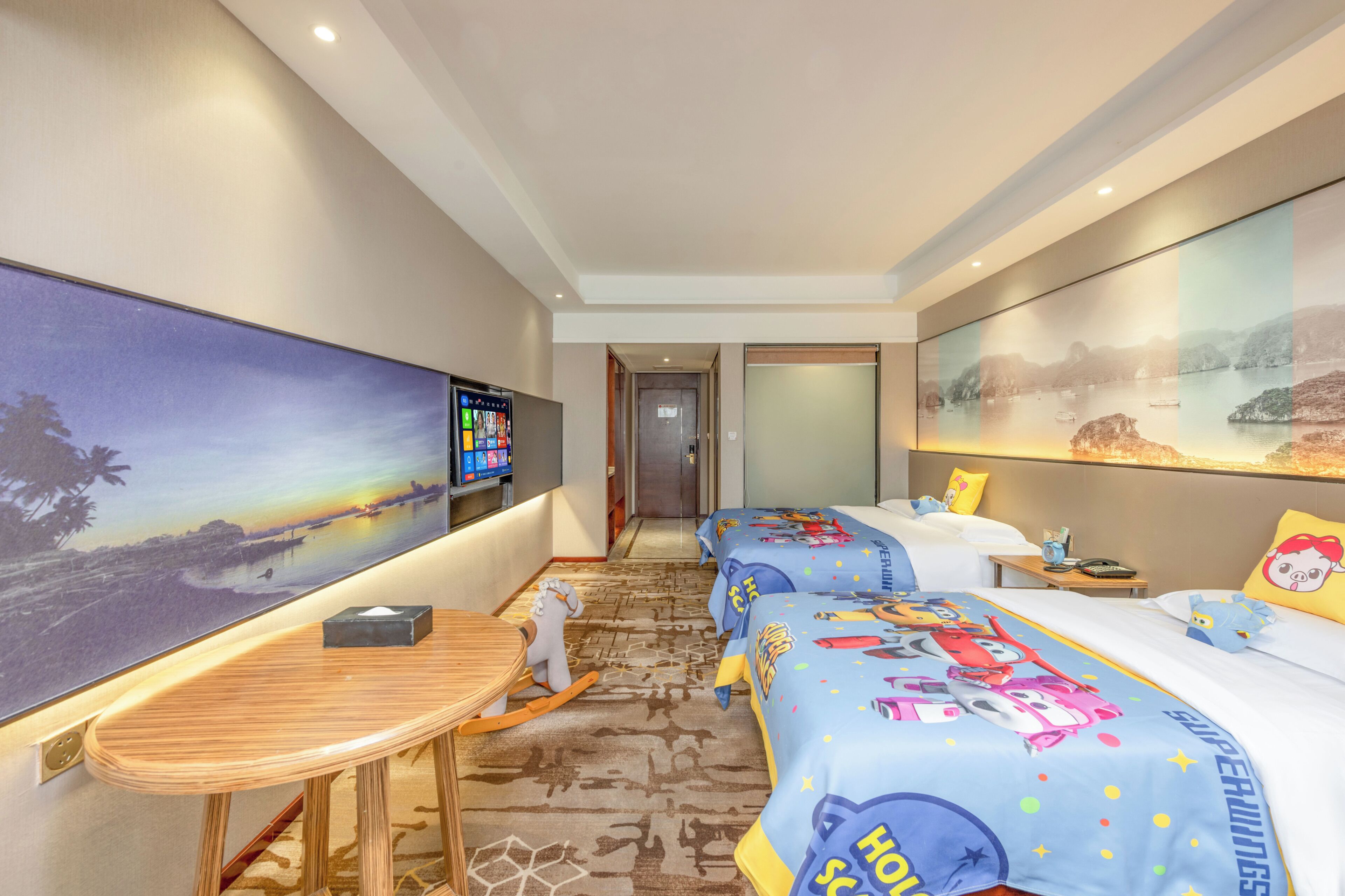 A hotel room with a large TV, a desk, two beds with colorful bedding, and a painting on the wall.