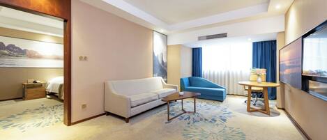 A hotel room with a sofa, a blue armchair, a small table, a TV, and a large window with curtains.
