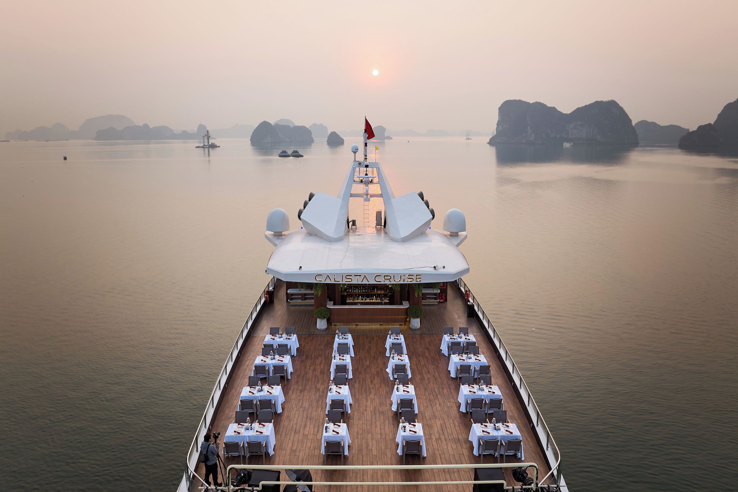 The deck of the Calista Cruise with tables and chairs arranged for dining, a flag at the stern, and distant islands in the background.