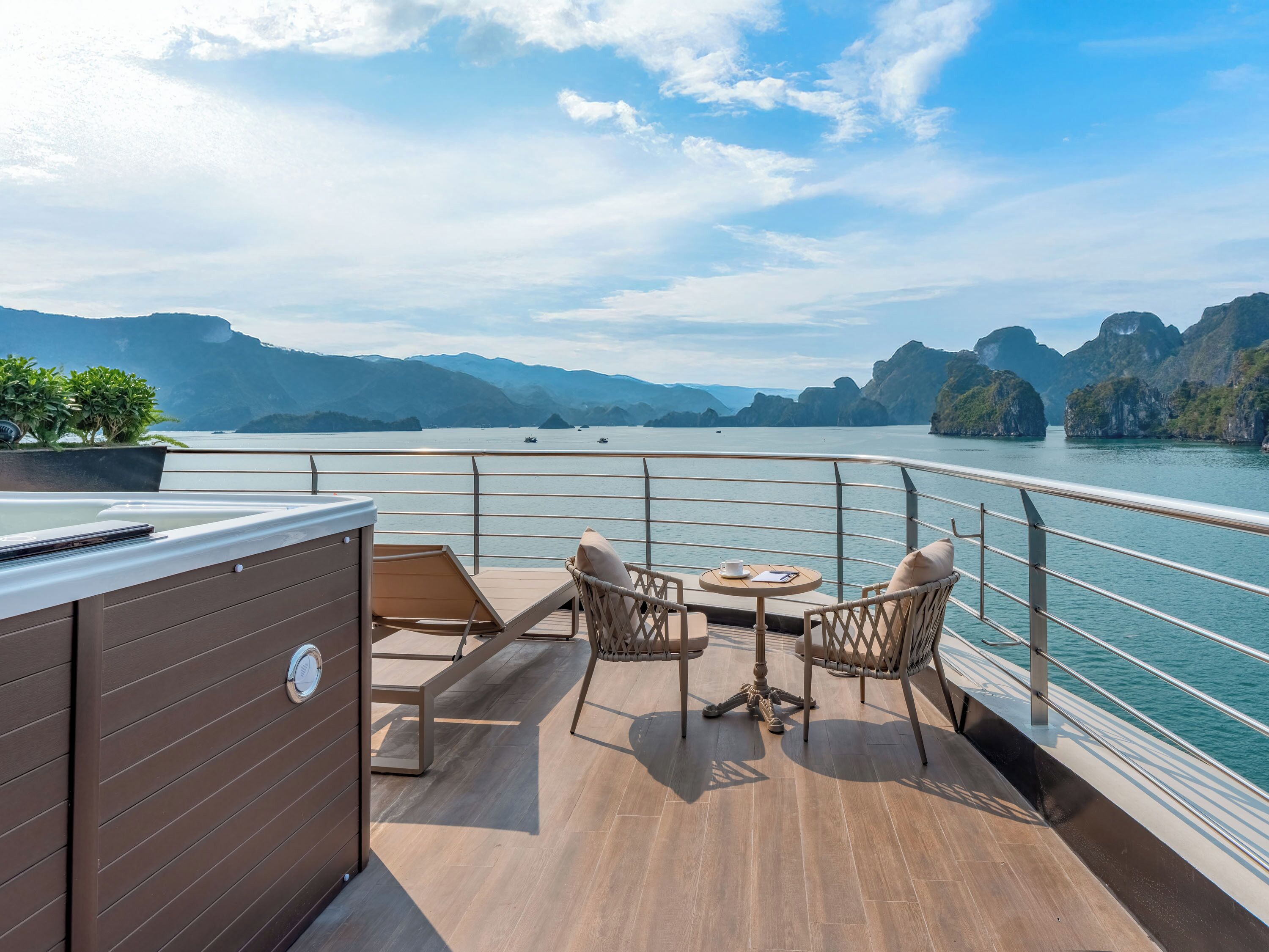 A deck with wooden flooring, a hot tub, and wicker chairs with a table, overlooking a calm lake and mountains.