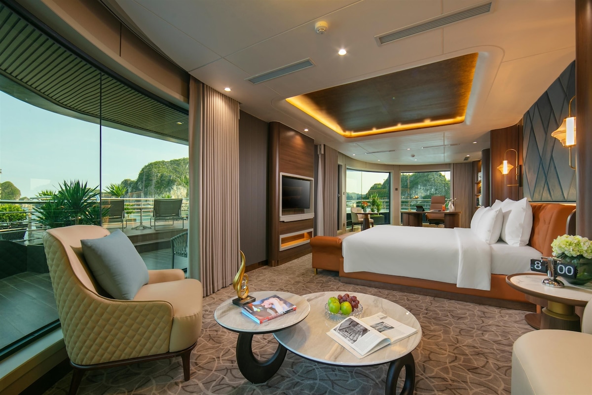 Una habitación de hotel moderna con una cama grande, televisión, zona de estar con una mesa y balcón con plantas.