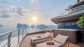 A sunlit deck with lounge chairs and a hot tub, overlooking a body of water and mountains. - Calista Cruise - A Ha Long Bay Spectacle (Tuan Chau)