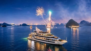 A large, multi-deck yacht with a modern design, sailing on calm waters at night with fireworks in the background.