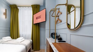 Une chambre d’hôtel avec un lit, une commode en bois, un miroir, un téléviseur affichant le message « CHAMBRE PRÊTE POUR L’ARRIVÉE DES CLIENTS » et une fenêtre avec des rideaux.