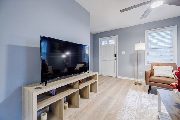 Una sala de estar con un televisor de pantalla plana sobre un soporte de madera, un sofá de cuero marrón, una lámpara de pie y una alfombra estampada.