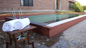 Una piscina con un banco de madera y toallas, un edificio de ladrillo al fondo y suelo de baldosas.