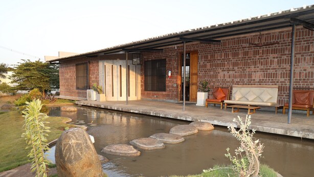 Una casa de ladrillo con porche cubierto, un pequeño estanque con piedras para cruzarlo y mobiliario de exterior.