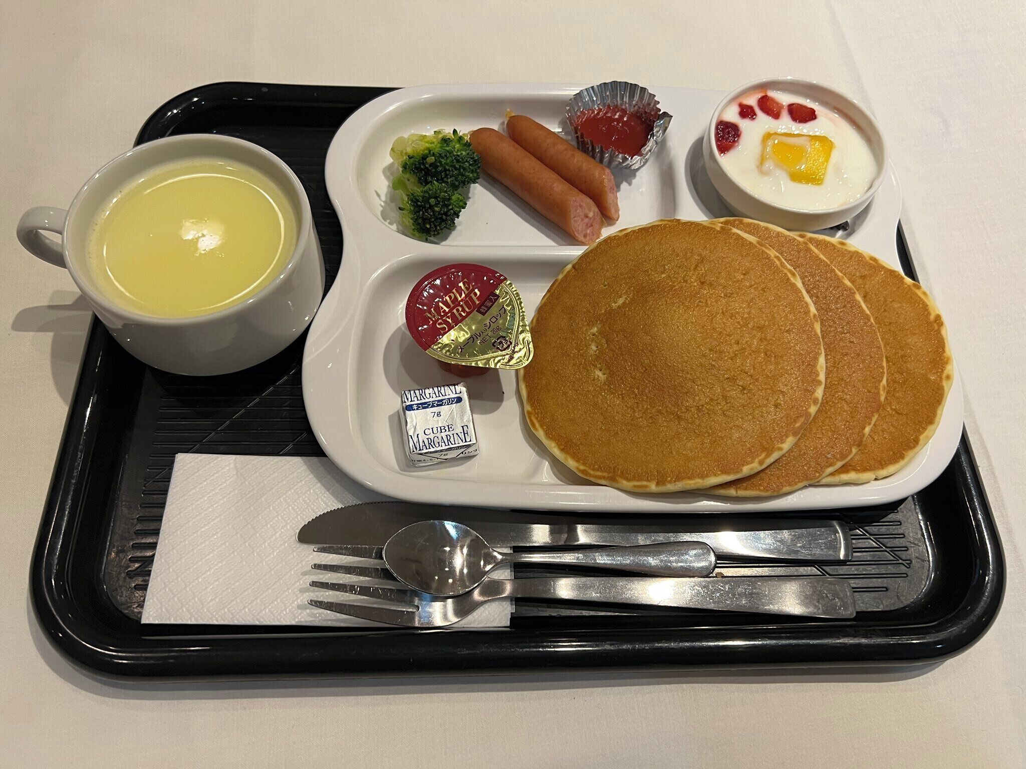 A tray with pancakes, hot dogs, broccoli, a cup of soup, and a small dish of fruit.