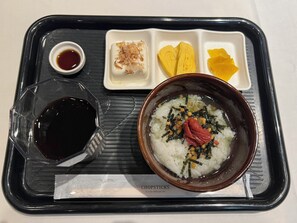 A meal tray with a bowl of rice topped with seaweed and red ingredients, a small dish of soy sauce, and a glass of dark liquid.
