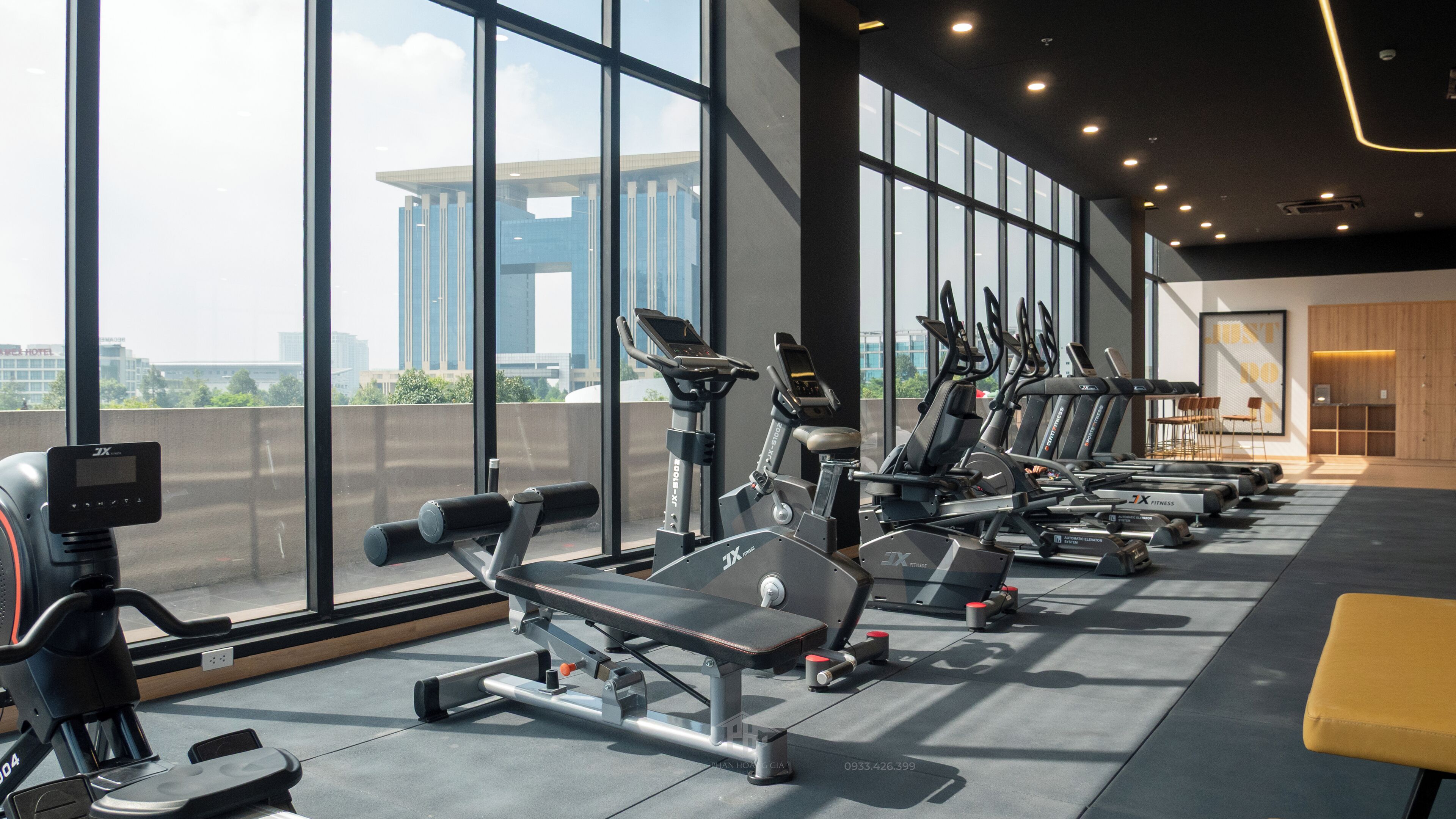 Een sportschool met cardioapparatuur en een bank, grote ramen en uitzicht op een gebouw.