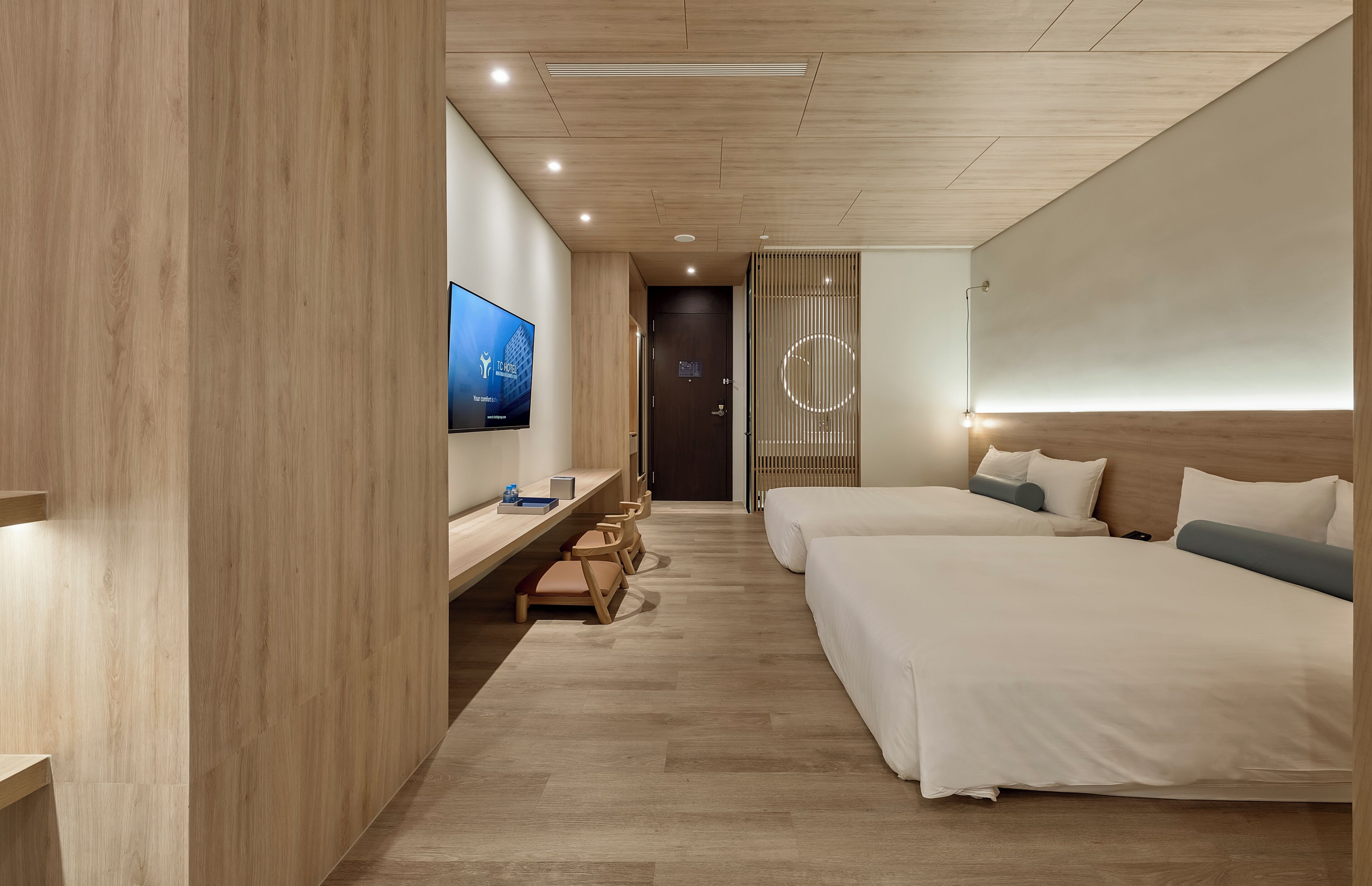 A modern hotel room with two beds, a desk with chairs, a TV, and a wooden floor.
