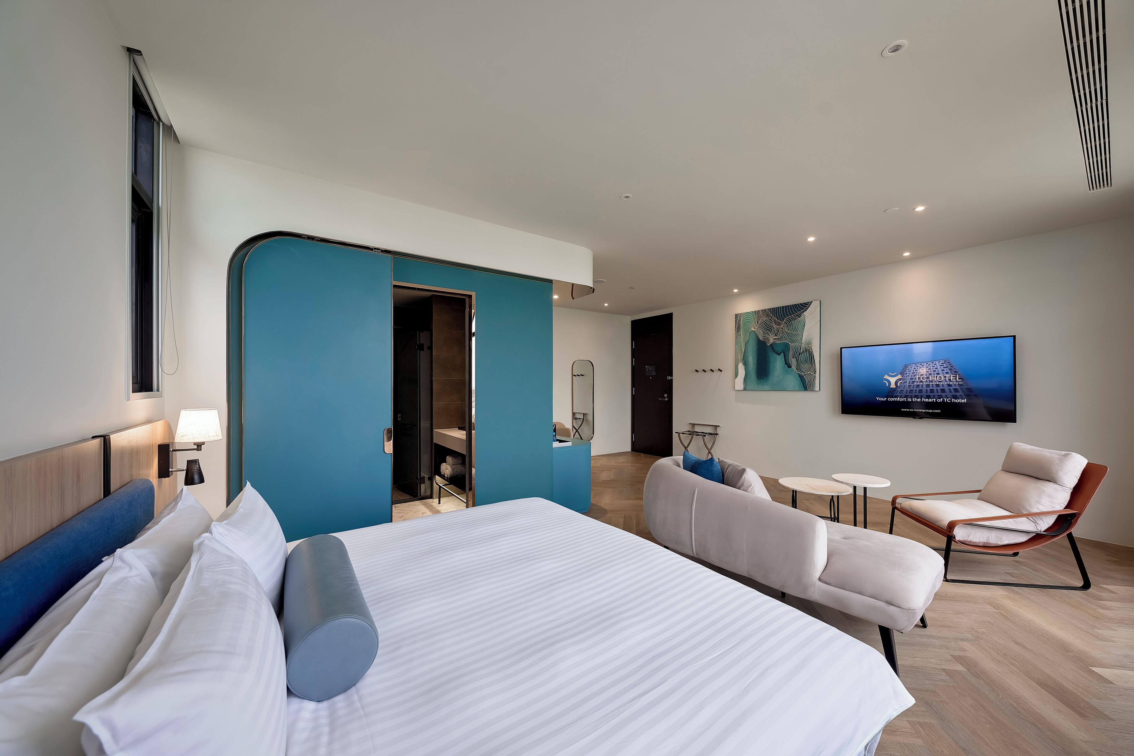 A modern hotel room with a large bed, a seating area with a sofa and chairs, a television, and a bathroom with a shower and a toilet.