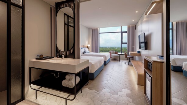 A modern hotel room with a large bed, a separate sofa, a desk, a bathroom with a large mirror, and a view of the outdoors.