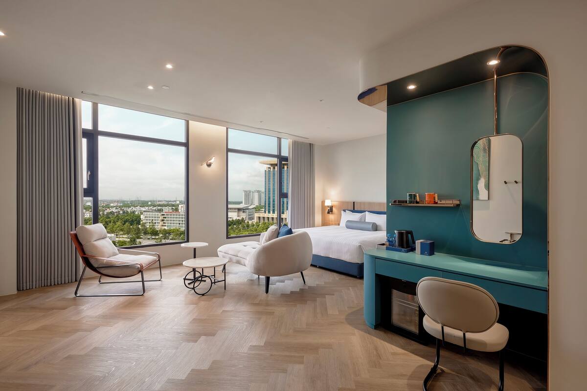 Une chambre d’hôtel moderne, dotée d’un grand lit, de deux fauteuils, d’une petite table ronde, d’une coiffeuse avec un miroir et offrant une vue sur le paysage urbain grâce à de grandes fenêtres.