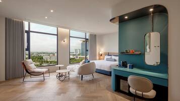 Une chambre d’hôtel moderne, dotée d’un grand lit, de deux fauteuils, d’une petite table ronde, d’une coiffeuse avec un miroir et offrant une vue sur le paysage urbain grâce à de grandes fenêtres.