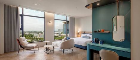 A modern hotel room with a large bed, two armchairs, a small round table, a vanity with a mirror, and a view of the cityscape through large windows.
