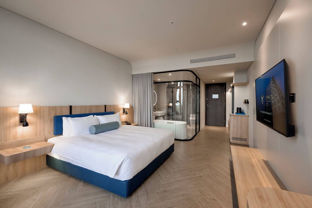 Une chambre d’hôtel moderne dotée d’un grand lit, de tables de chevet, d’une salle de bain fermée par des parois de verre et d’un téléviseur fixé au mur.