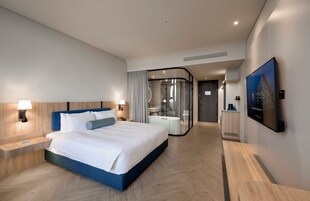 A modern hotel room with a large bed, bedside tables, a glass-enclosed bathroom, and a TV mounted on the wall.