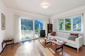 A spacious living room with a white sofa, a coffee table, and a bookshelf. Large windows offer a view of a mountainous landscape.
