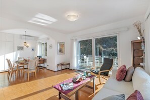 A spacious living area with a dining table set for four, a sofa with cushions, and a large window offering a view of the outdoors. - Castel In Aria 2 - Val Verzasca, Vogorno, Switzerland (Vogorno)