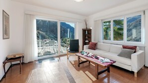 A spacious living room with a white sofa, a wooden coffee table, and a large window offering a mountain view.