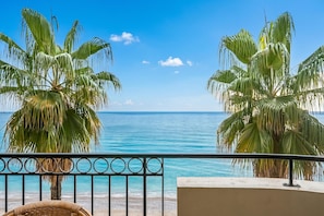 Un balcón con vista al mar, palmeras y una silla de mimbre.