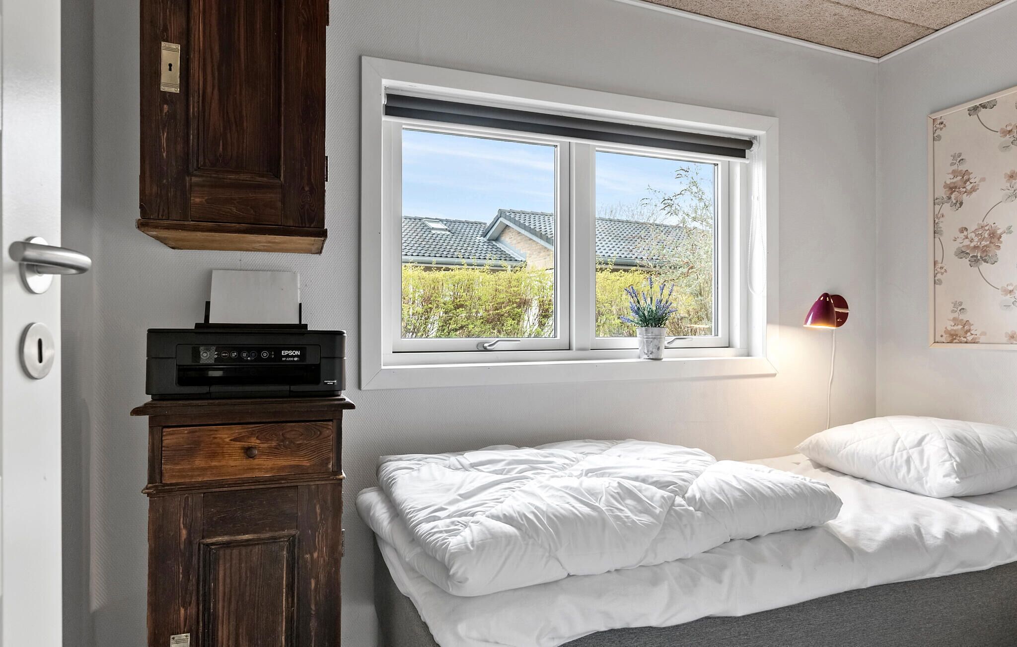 A bedroom with a bed, a window with a view of houses, a desk with a printer, and a wall-mounted lamp.
