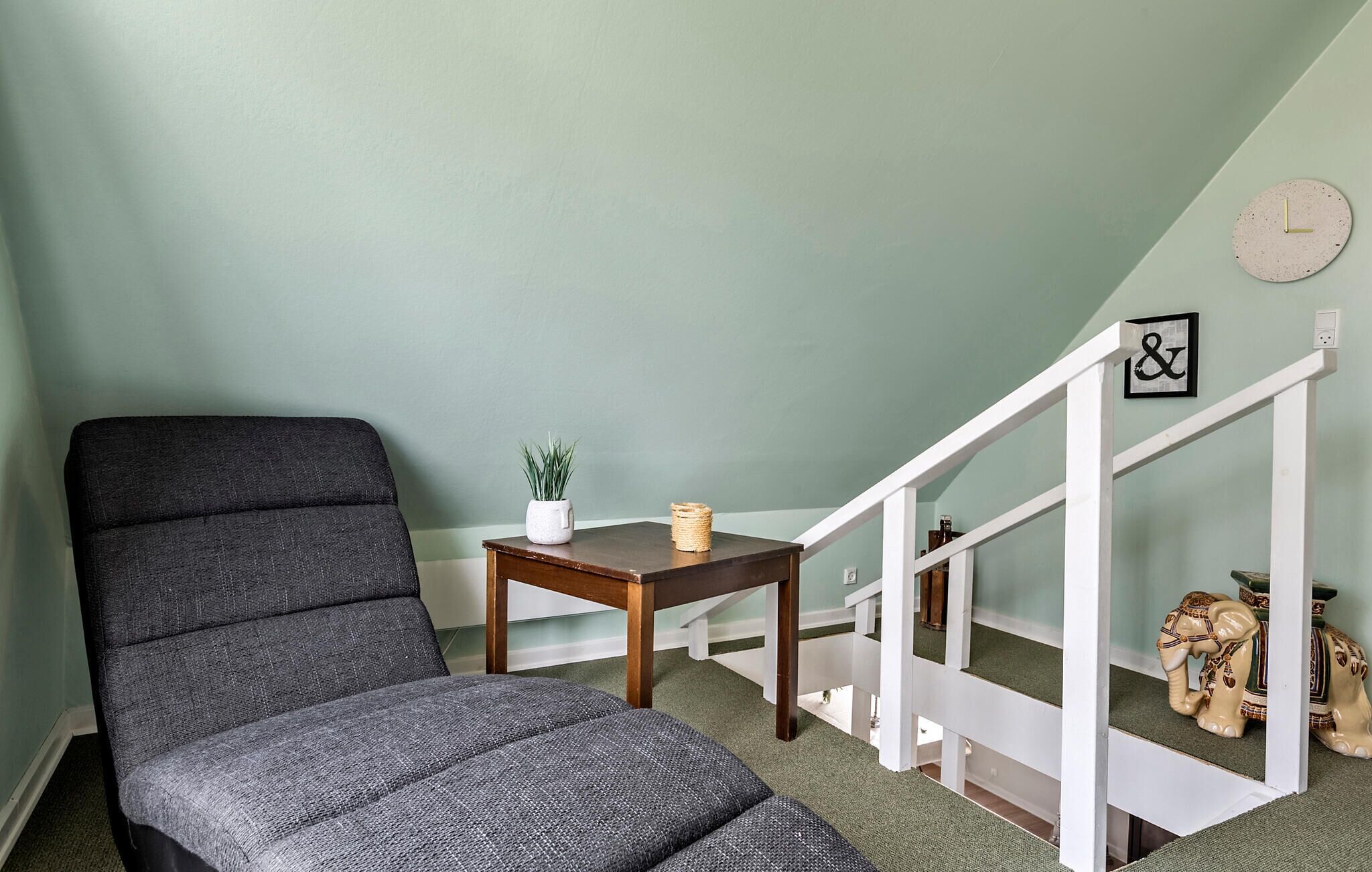 Habitación de esquina con una butaca tapizada de gris, una pequeña mesa de madera con una planta y una escalera blanca con un reloj y un cuadro enmarcado.