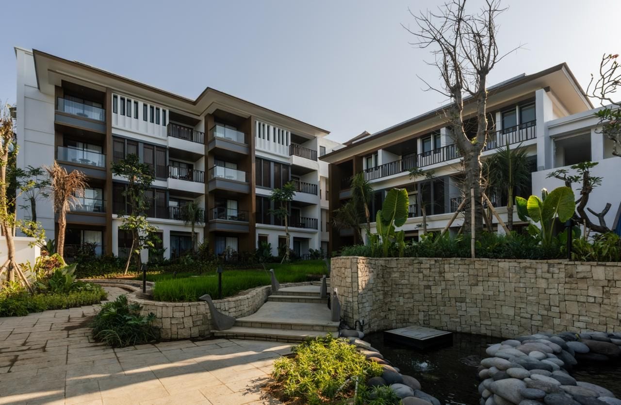 A multi-story hotel with balconies, a landscaped garden, and a small water feature.