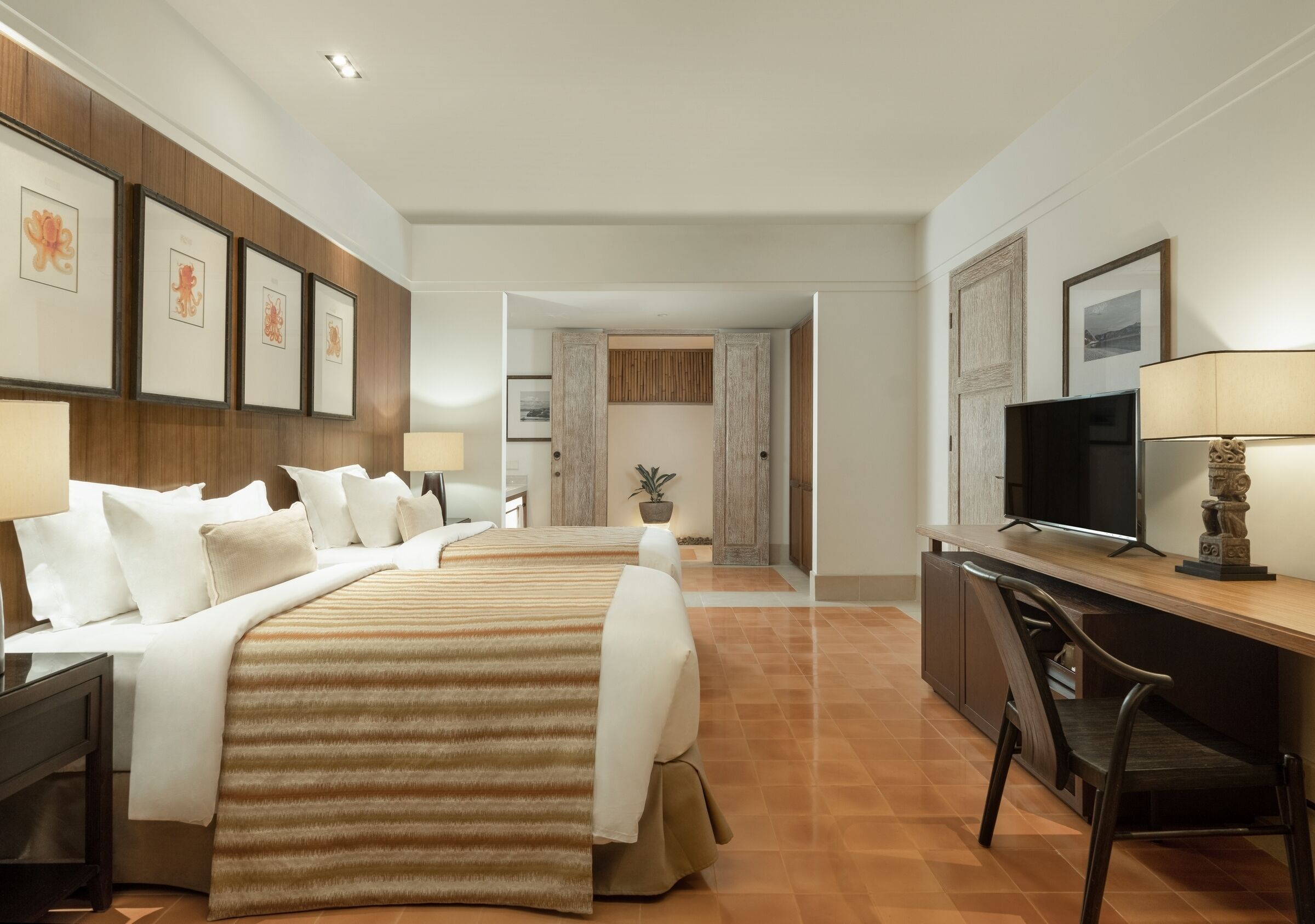 A hotel room with two beds, a desk, a TV, and framed artwork on the walls.