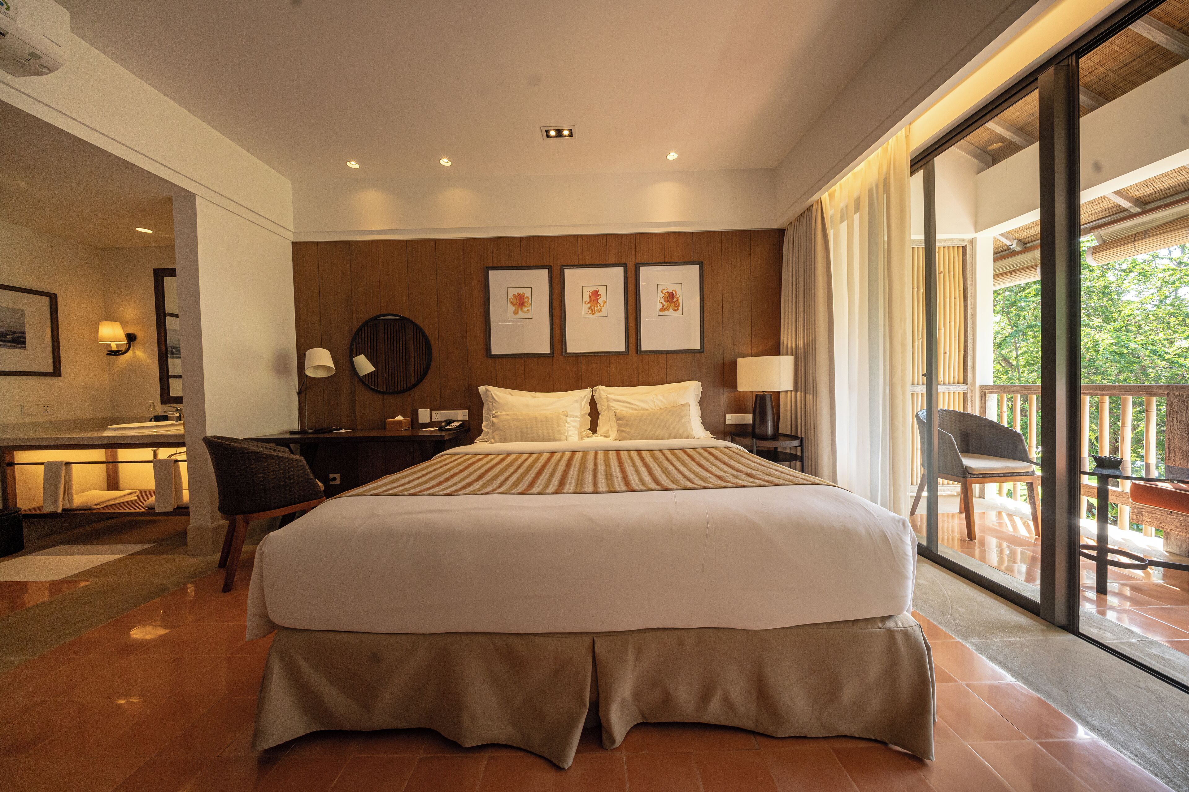 A spacious bedroom with a large bed, a desk, and a chair, a wooden headboard, and a balcony with a view of trees.