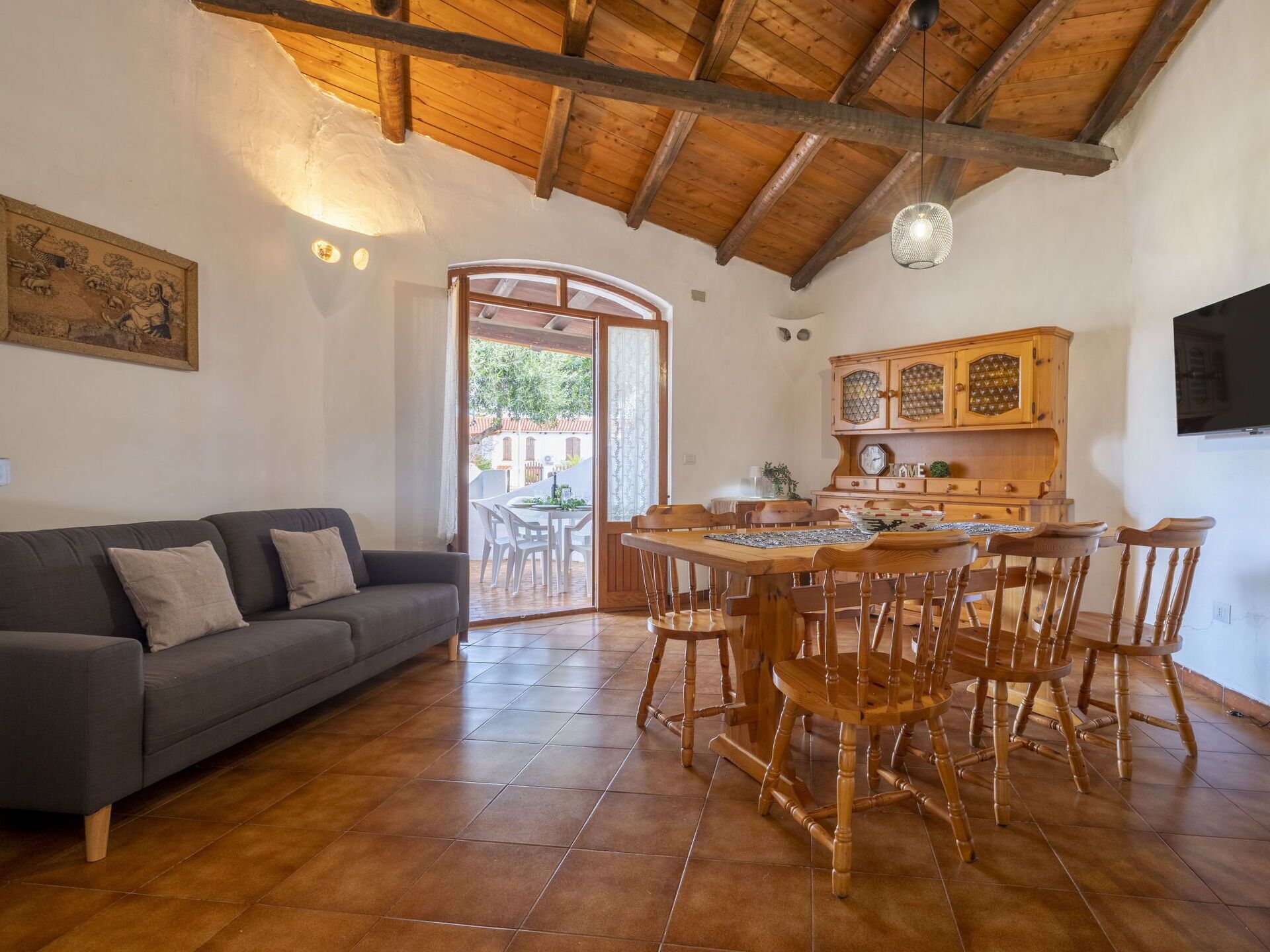 A cozy living room with a wooden ceiling, a sofa, a dining table with chairs, and a cabinet with a television.