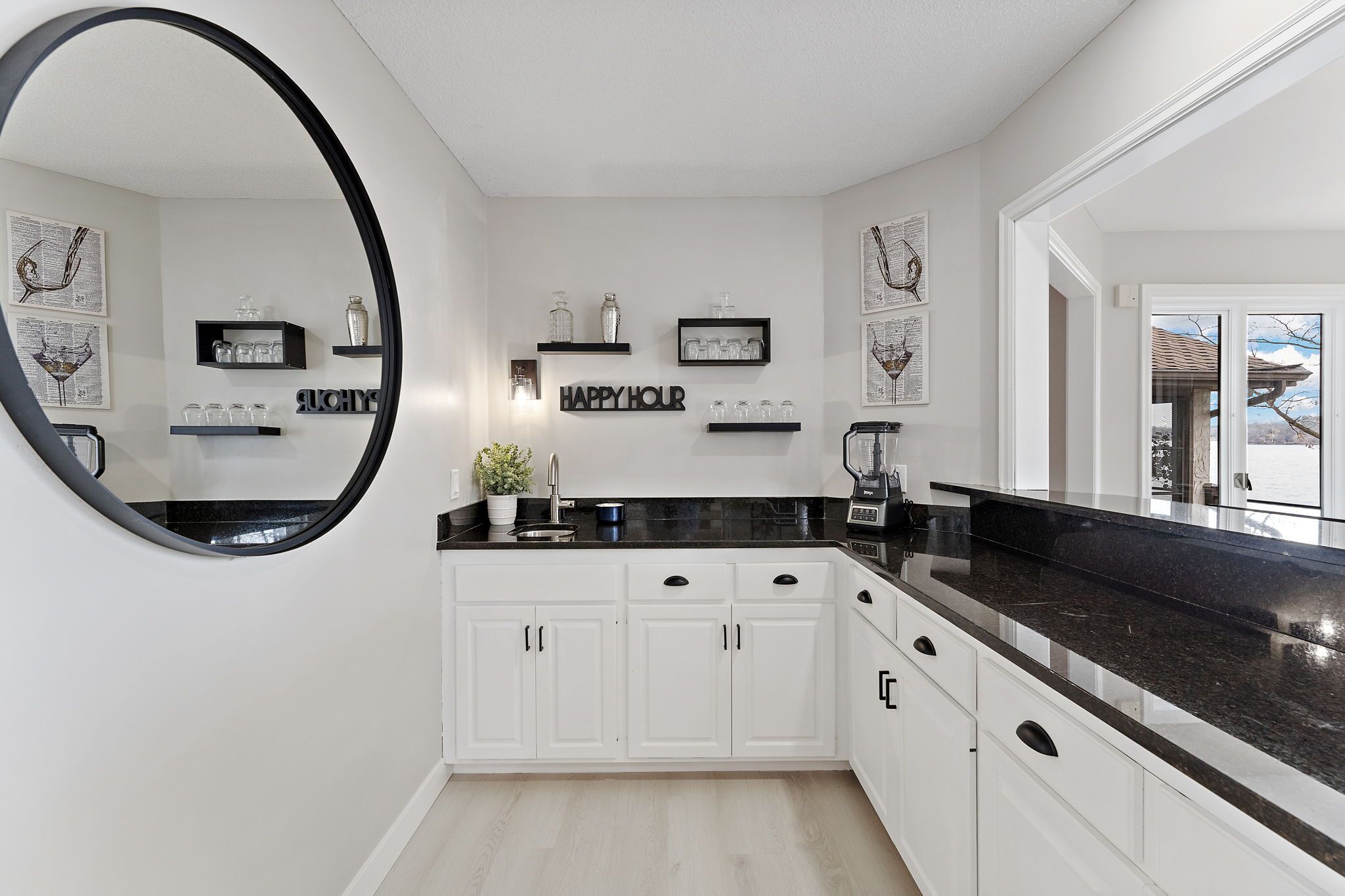 A modern kitchen with white cabinets, black countertops, and a large mirror. There are decorative items on shelves and a 'Happy Hour' sign.