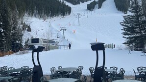 Zona exterior con mesas y sillas cubierta de nieve, con vista a una pista de esquí que incluye un telesilla y una persona caminando.