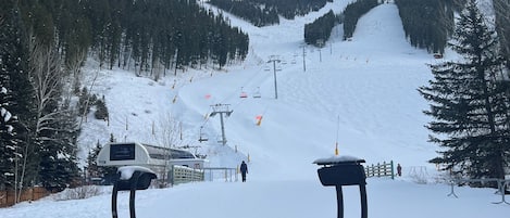 A snow-covered outdoor seating area with tables and chairs, overlooking a ski slope with a chairlift and a person walking.