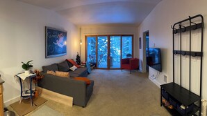 Una sala de estar con un sofá gris, una pequeña mesita de noche con una planta, un sillón rojo, un televisor y un ventanal corredero que da a un balcón.