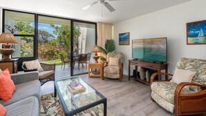 A living room with a glass coffee table, a sofa, a chair, a television, and a ceiling fan. - Lush 1BD garden view, Steps from Beach & Pool - Maui Vista 1106 (Kihei)