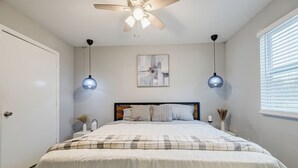 Un dormitorio con una cama, un ventilador de techo, dos lámparas colgantes, un cuadro abstracto en la pared y una ventana con persianas.