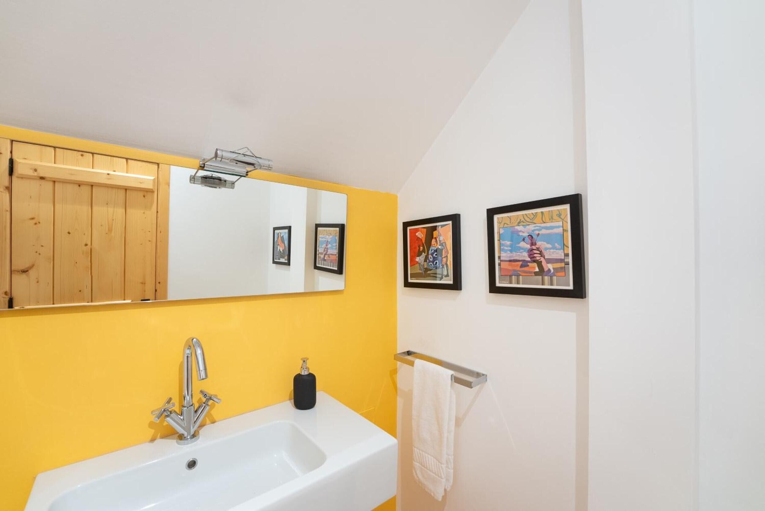 Une salle de bain avec un lavabo blanc, des murs jaunes, un panneau en bois, un miroir et deux tableaux encadrés.
