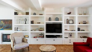 A living room with a TV, shelves, a fireplace, a red sofa, and a beige armchair.