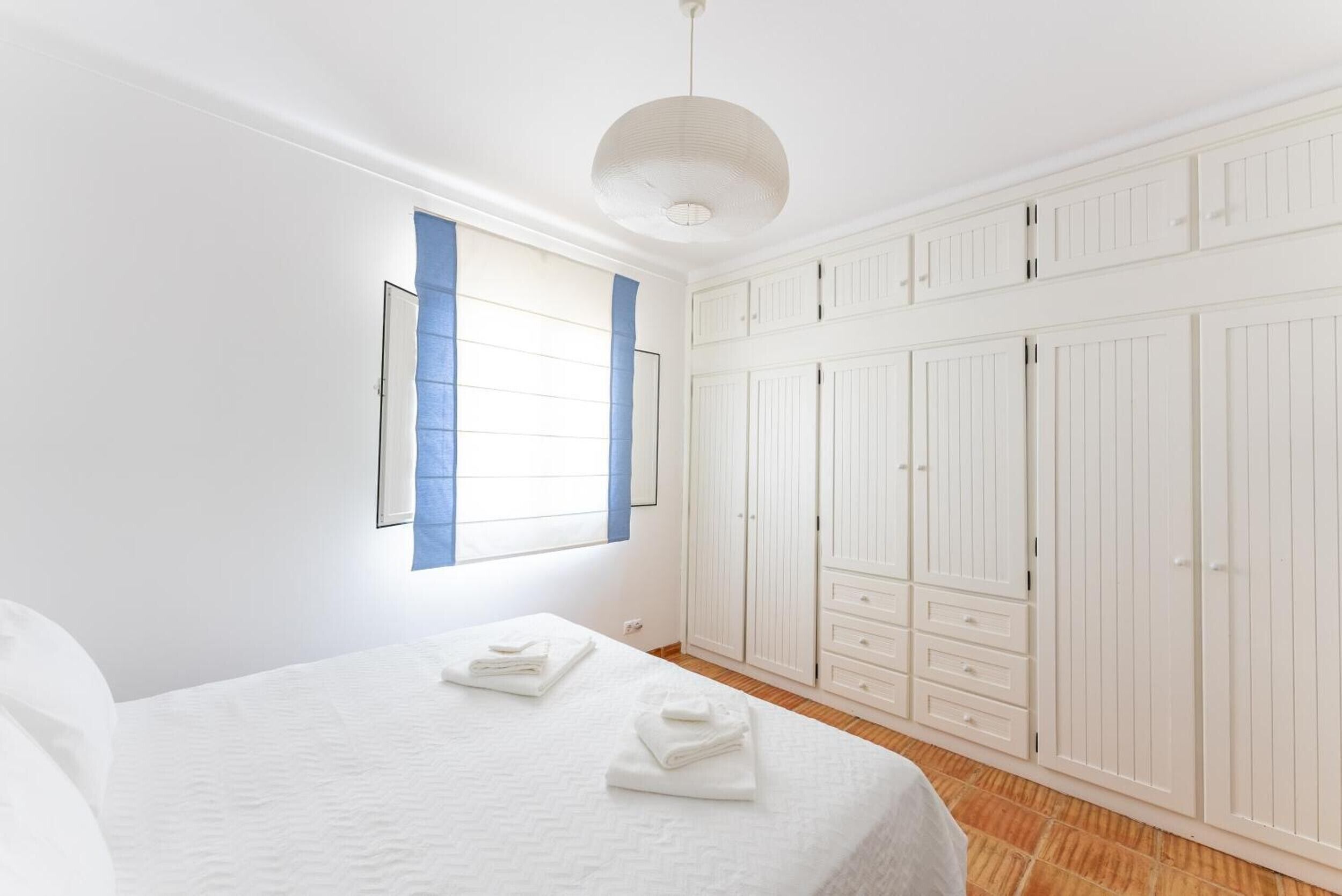 A bedroom with a bed, a wardrobe, a window with blinds, and a hanging light fixture.