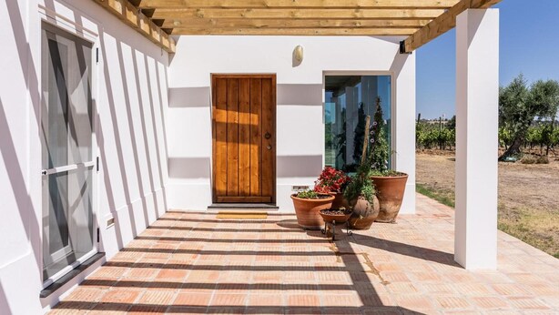 A covered outdoor area with a wooden pergola, a white building with a wooden door, and potted plants.