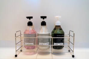 Estante metálico con tres botellas de jabón líquido etiquetadas como "champú", "acondicionador" y "gel de baño".