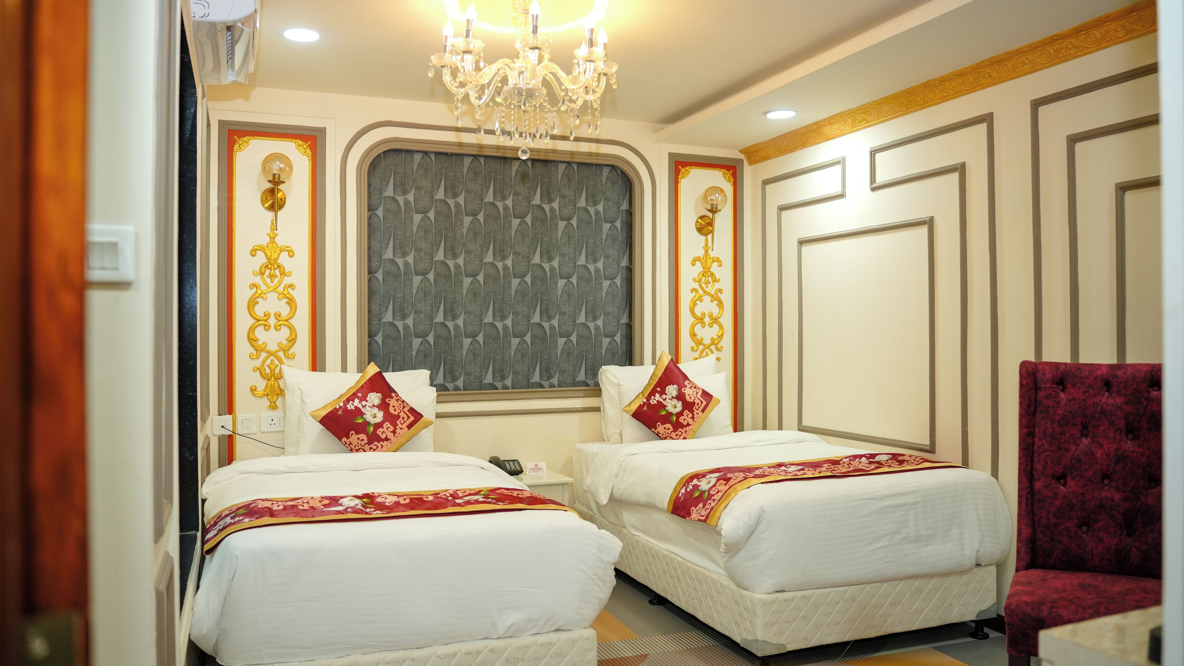 Habitación de hotel con dos camas, un sillón de terciopelo rojo, una araña y paneles decorativos en la pared.
