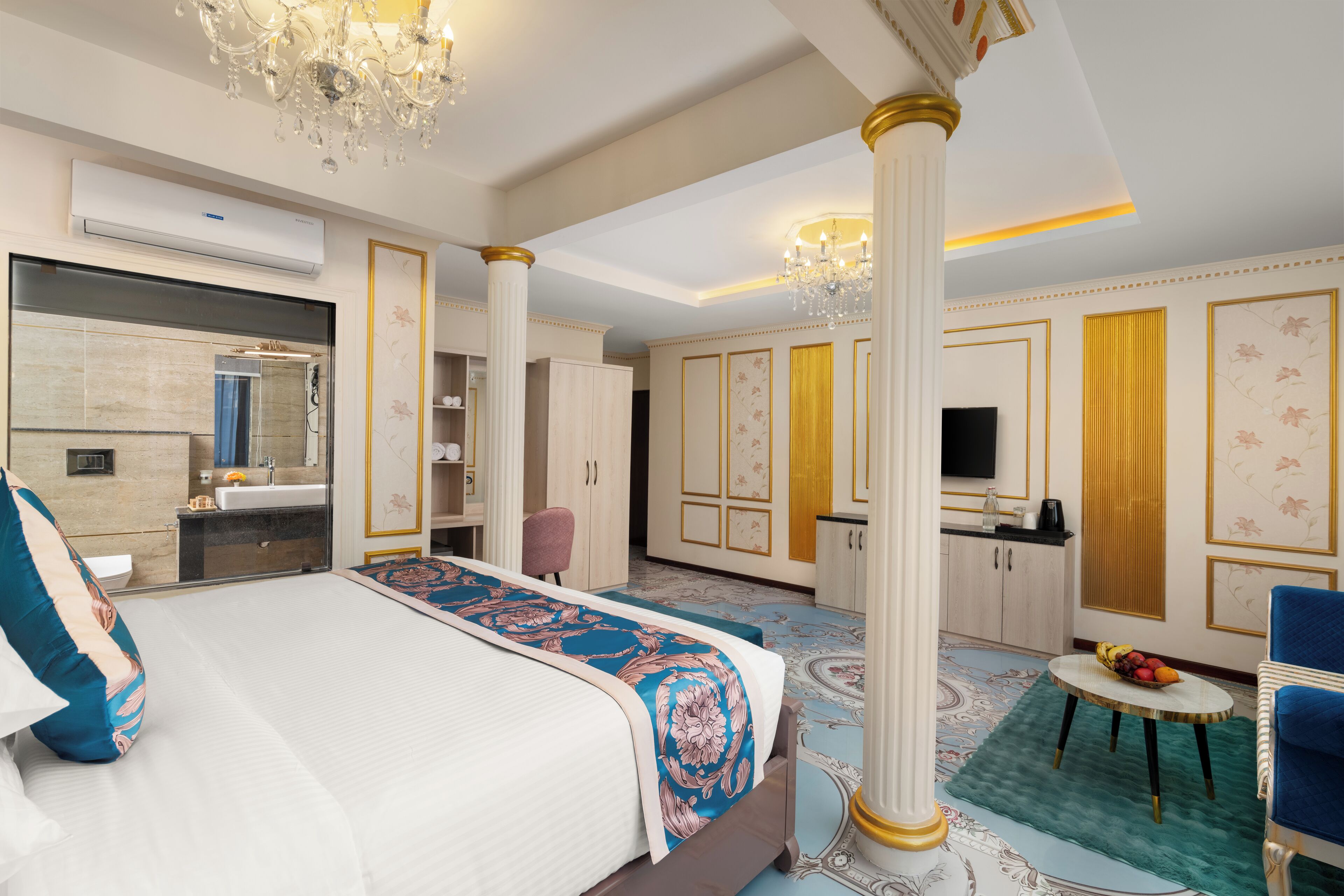 A spacious bedroom with a large bed, a sitting area with a table and chairs, a vanity with a mirror, and a small kitchen area.