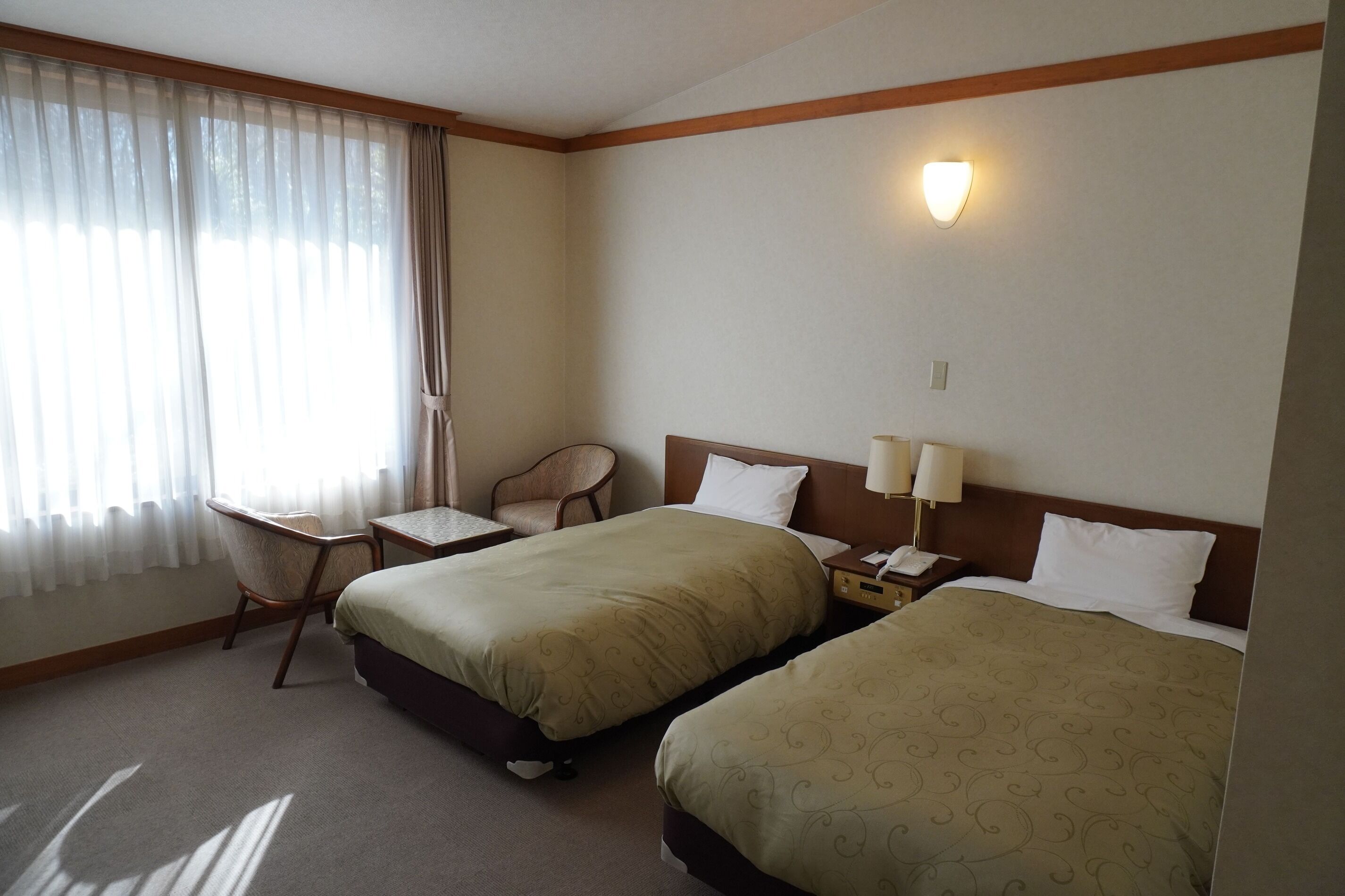Una camera d'albergo con due letti, una sedia, un tavolino e un comodino con una lampada.