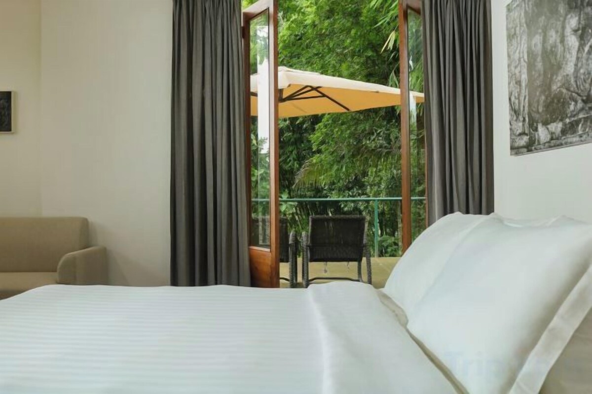 A hotel room with a bed, a chair, and a balcony with a view of greenery.