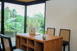 Une table en bois, sur laquelle se trouvent un ordinateur portable et un vase avec une plante, est entourée de chaises, dans une pièce dotée de grandes fenêtres donnant sur un espace verdoyant.